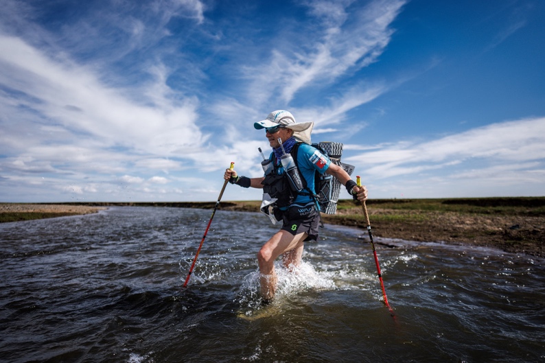 Roberto Rivola wading through a river with trekking poles and a loaded pack under a wide blue sky during the Gobi March in Mongolia
